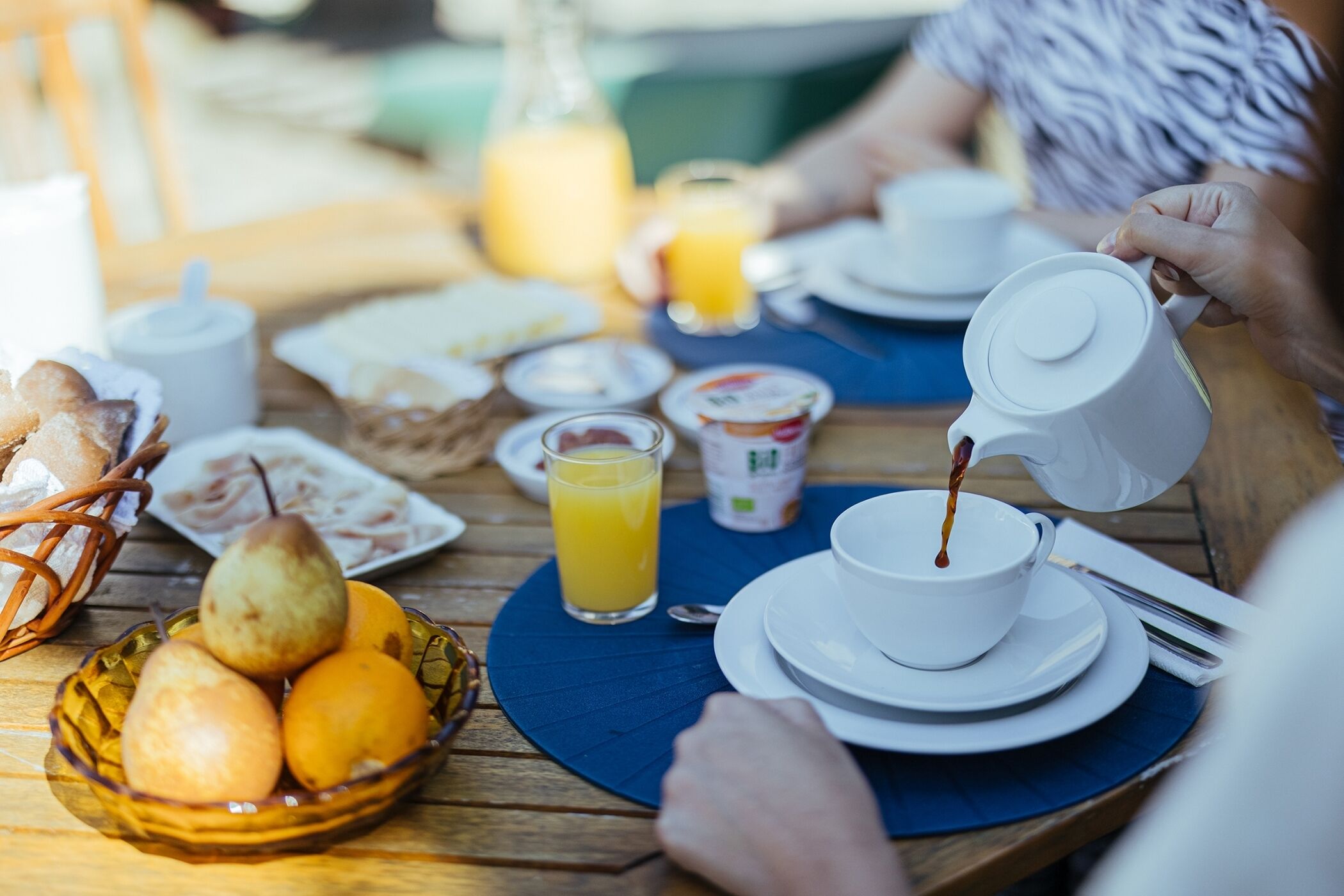 A breakfast table with a bowl of fruit, a glass of orange juice, and a white teapot pouring coffee.