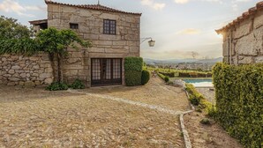 A stone building with a tiled roof, a door, and a window. There is a stone wall, a small garden, and a pool in the background.