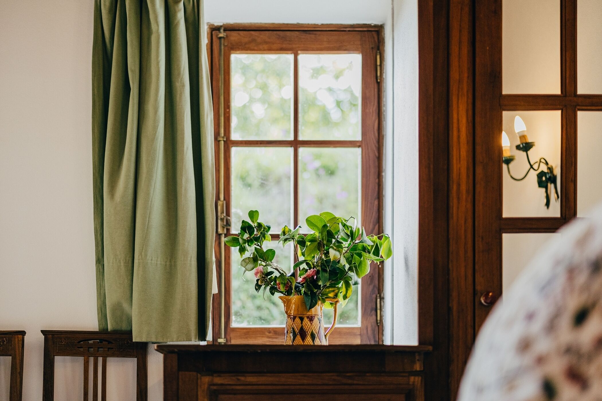 Une fenêtre avec un cadre en bois, un rideau vert et un vase avec des plantes sur une table en bois.
