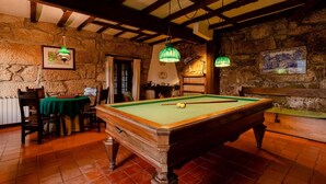 Una sala de billar con mesa de billar, vigas de madera, paredes de piedra y lámparas verdes colgantes.