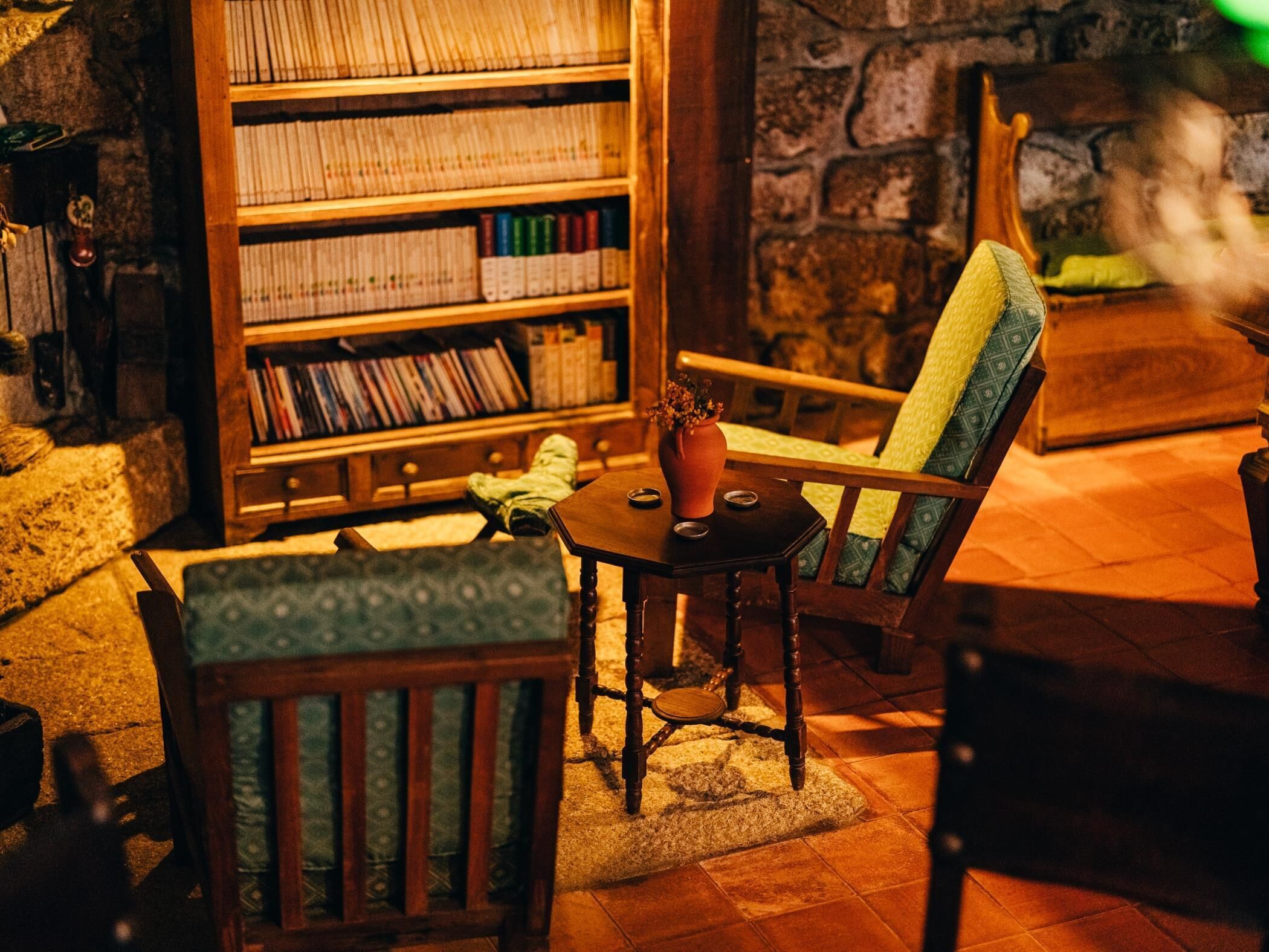 A cozy reading nook with a wooden bookshelf, a green upholstered chair, and a small table with a vase.