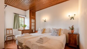 Un dormitorio con dos camas, techo de madera, ventana con cortinas, mesita de noche con una planta y una lámpara fijada a la pared.