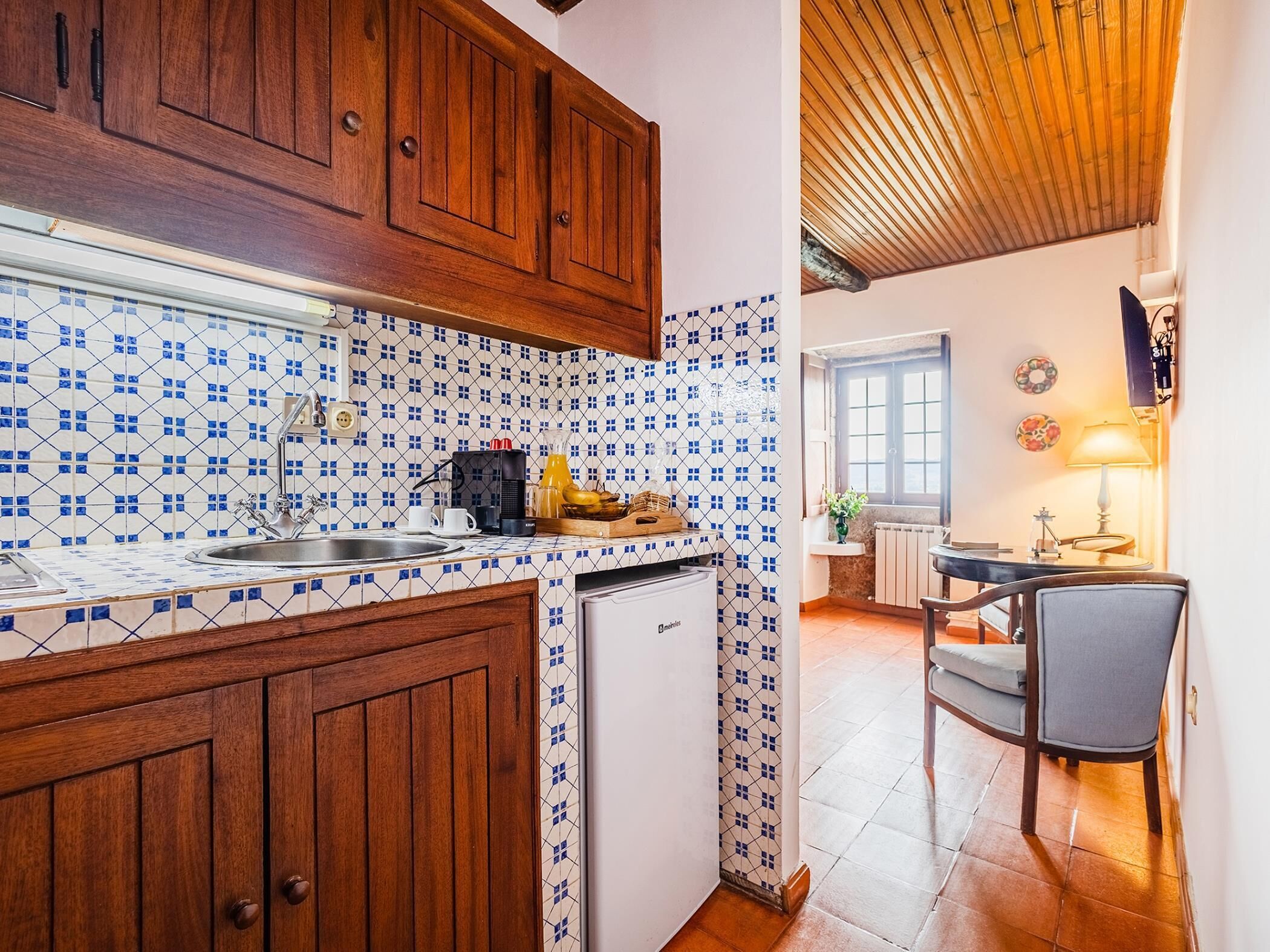 Een kleine keuken met houten kastjes, een betegelde spatwand en een compacte koelkast.