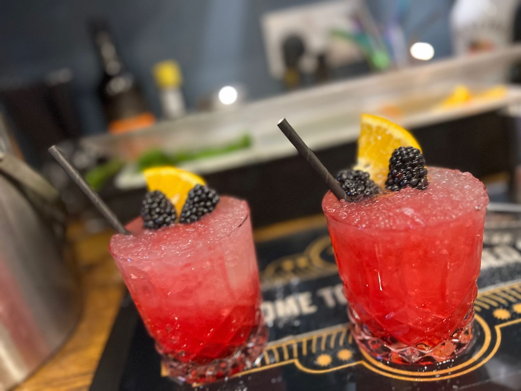 Dois cocktails com amoras e uma fatia de laranja como guarnição, servidos em copos transparentes sobre um apoio com um logotipo.