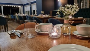 Una mesa de comedor dispuesta con un plato blanco, una taza rosa, una copa de vidrio transparente y otra copa de vidrio con una vela. En la mesa hay un jarrón pequeño con flores blancas.