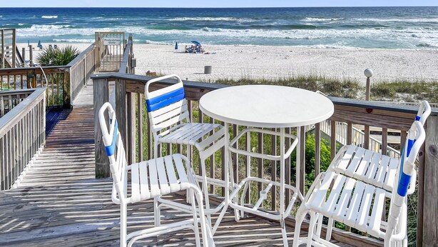 Une terrasse en bois avec des chaises et une table en métal peintes en blanc, donnant sur une plage où l’on peut admirer les vagues et un ciel dégagé.