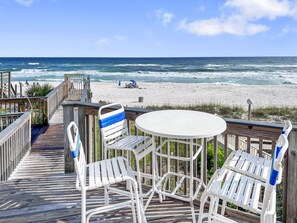 A wooden deck with white metal chairs and a table, overlooking a beach with waves and a clear sky. - Come Be At Peace! Spacious 3 Bd/3ba Beach Home! Sleeps 10! (Panama City Beach)