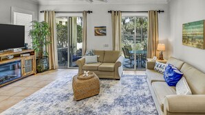 Un salon avec un canapé beige, un téléviseur, une table basse et une grande fenêtre avec des rideaux.