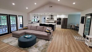 A spacious living area with a large L-shaped sectional sofa, a round ottoman, and a dining table with chairs. The room features a kitchen with modern appliances and pendant lighting. - Mid-century Modern Remodel near Pisgah & Asheville (Asheville)