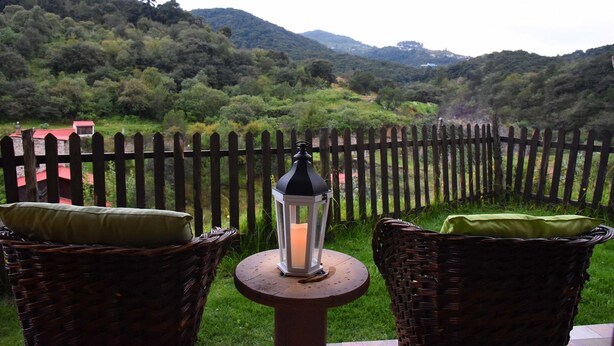 A wicker chair with a cushion and a lantern on a table, overlooking a fenced garden with a view of hills. - Cabaña El Encino en Real del Monte (Mineral del Monte)