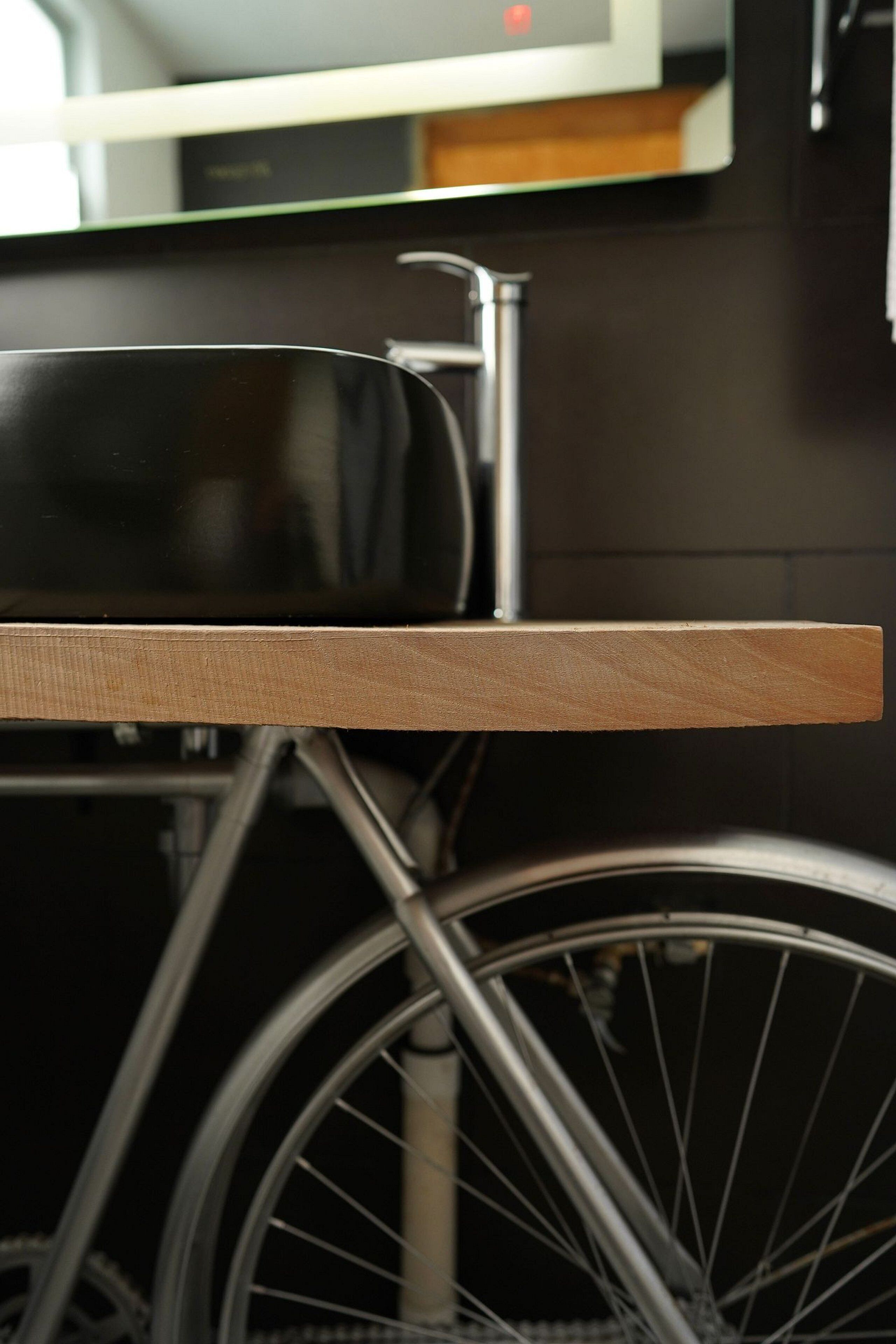 A wooden table with a black chair, a bicycle wheel, and a metal structure in the background.