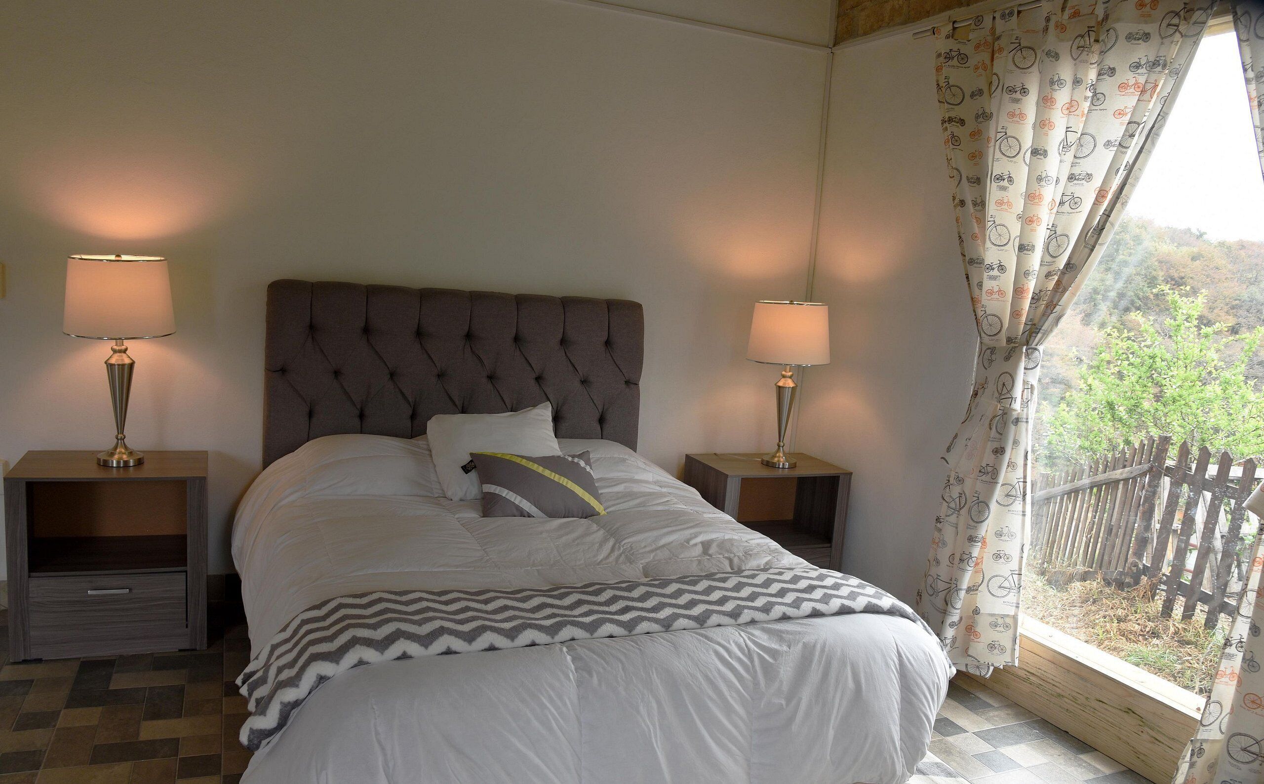 A bedroom with a tufted headboard, two bedside tables with lamps, a bed with white bedding and a patterned throw, and a window with curtains.