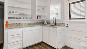 Una cocina con gabinetes blancos, encimera de granito, un lavamanos y una ventana con persianas.