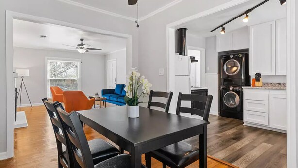 Una cocina moderna con lavadora y secadora integradas, un comedor con mesa y sillas oscuras, y una sala de estar con un sofá azul y sillas naranjas.