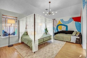 A bedroom with a four-poster bed, a bedside table, a window with curtains, and a wall with abstract art.