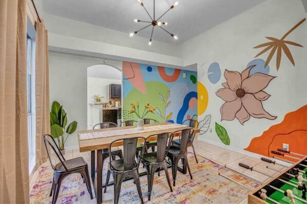 A dining area with a wooden table and chairs, a colorful abstract mural on the wall, and a foosball table.