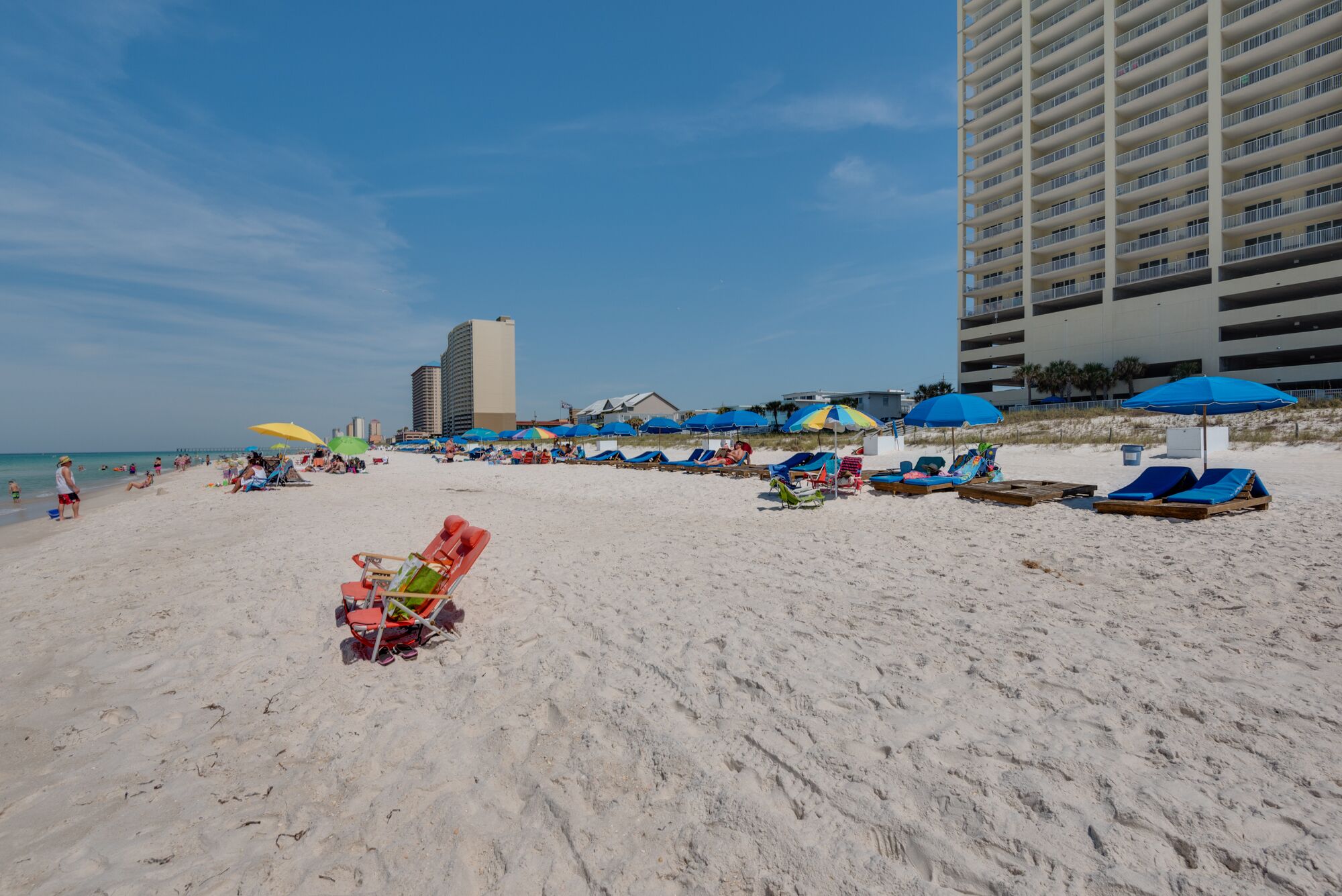 A beach with sun loungers and umbrellas, a tall building in the background, and a clear blue sky.