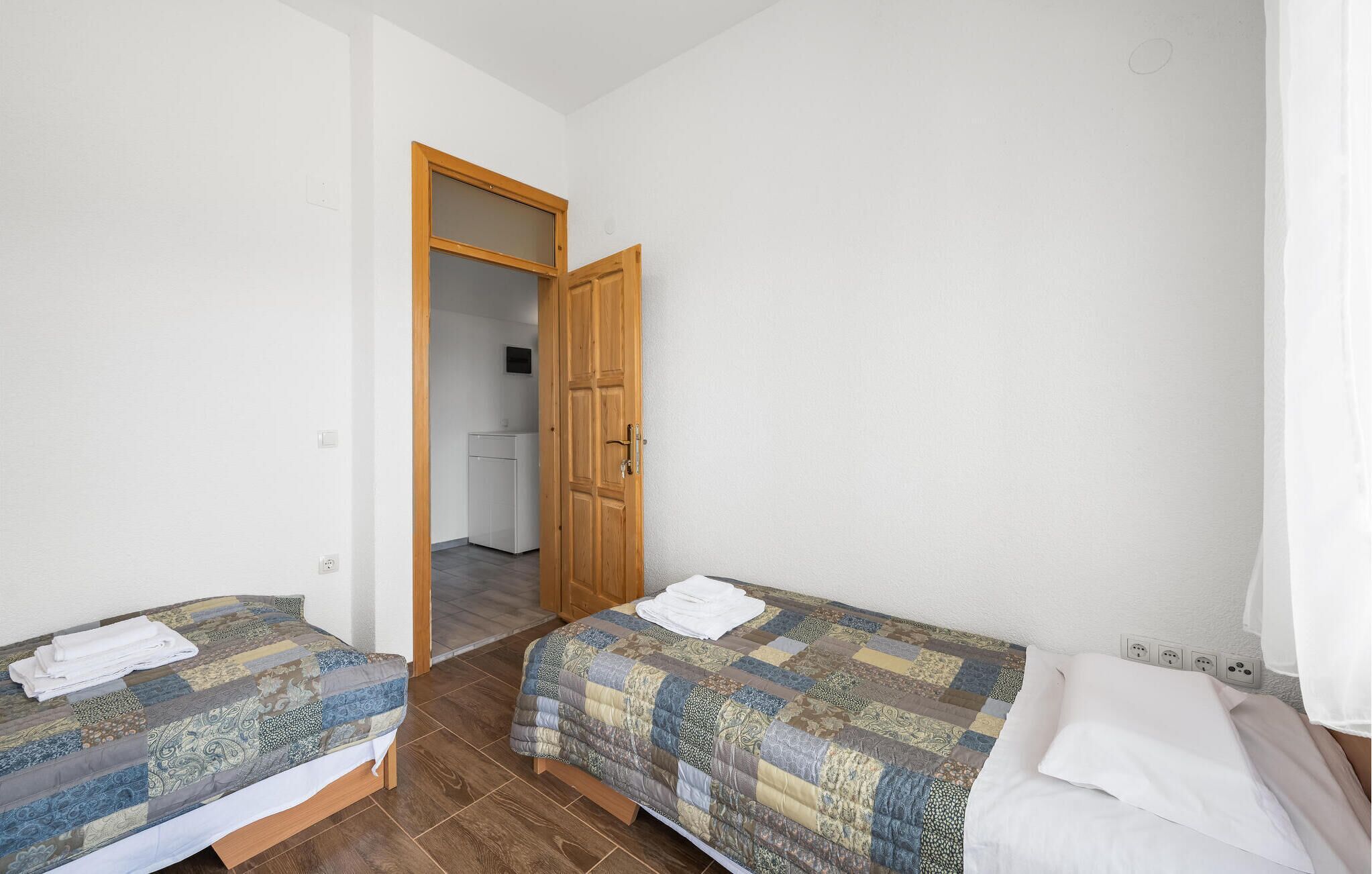 A small, clean room with two single beds, a wooden door, and a kitchenette in the background.