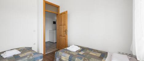A small, clean room with two single beds, a wooden door, and a kitchenette in the background.