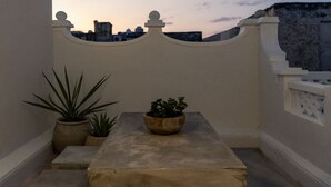 A balcony with a table and potted plants, a patterned floor, and a cityscape view at dusk.