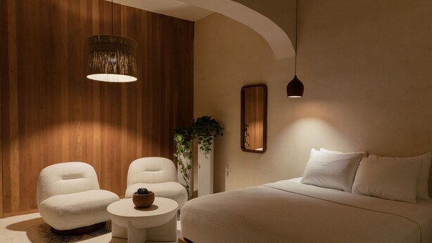 A modern bedroom with a large bed, two white armchairs, a small round table, and a hanging light fixture.