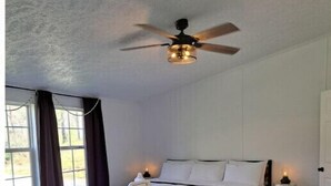 A bedroom with a bed, a ceiling fan, a bedside table with a lamp, and a window with curtains. - 50 acre homestead to hike, forage, harvest, explore, & ENJOY! (Sistersville)