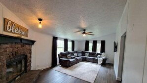 A spacious living room with a stone fireplace, a sectional sofa, and a ceiling fan. - 50 acre homestead to hike, forage, harvest, explore, & ENJOY! (Sistersville)