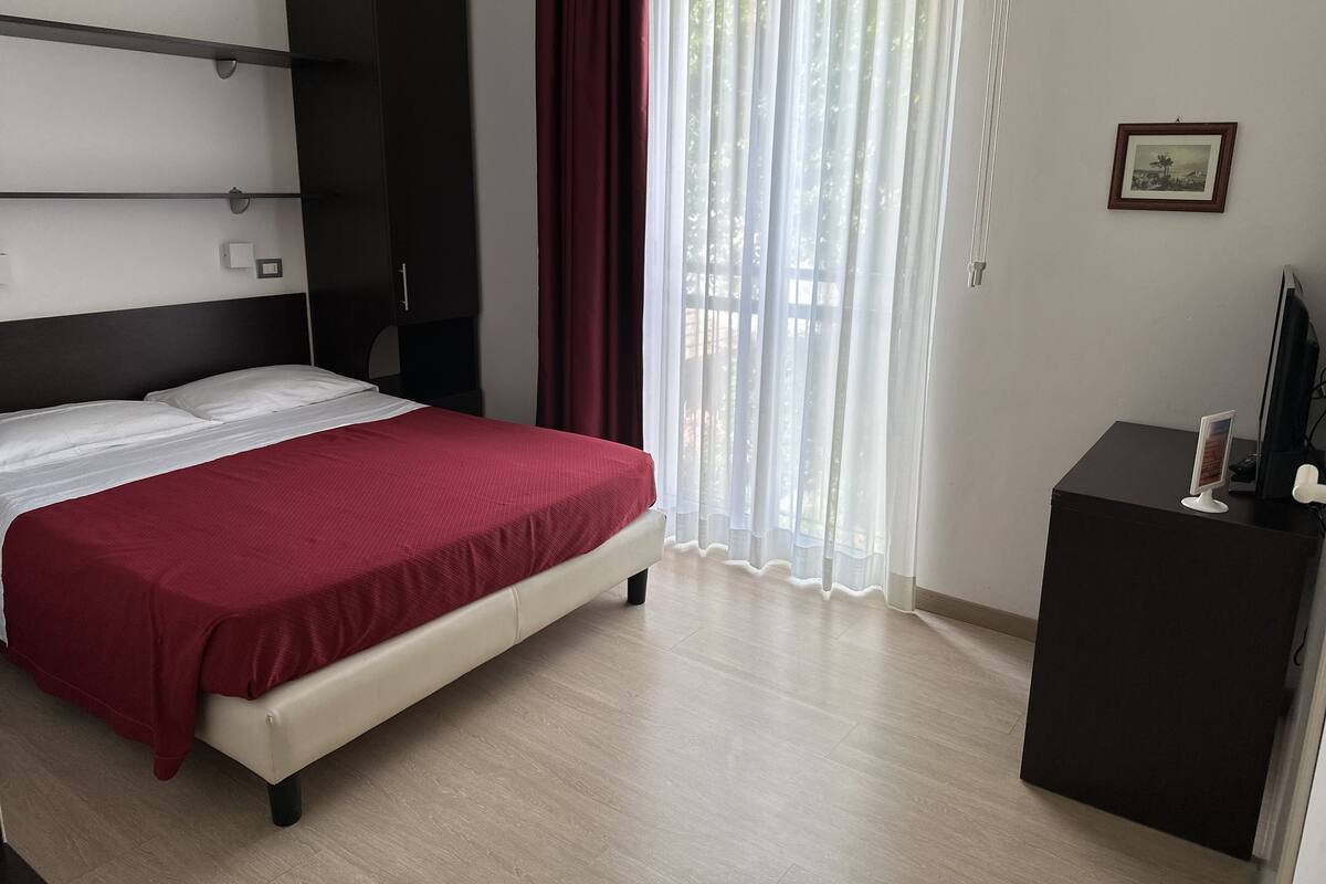 A hotel room with a bed, a wardrobe, a desk with a computer, and a window with curtains.