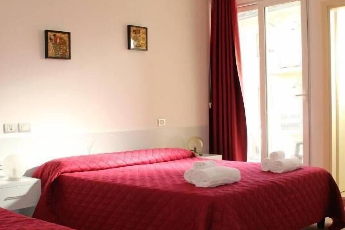 A hotel room with two beds, a window with red curtains, and a view of the outside.
