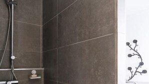 Un espacio de ducha con cabezal de ducha de mano, panel de control en la pared y un estante pequeño con artículos de aseo.