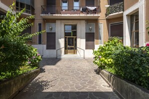 A courtyard with a stone pathway leading to a building entrance, surrounded by greenery and a balcony with a railing.
