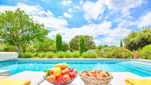 A poolside setting with a table holding fruit bowls and a wicker basket, surrounded by lush greenery and a clear blue sky. - Mas des Chênes Verts – 5 ch avec piscine et clim (Eygalières)