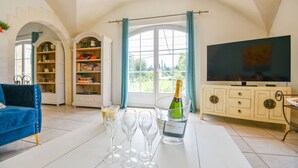 A living room with a blue sofa, a bookshelf, a TV, and a table with wine glasses and a bottle of champagne. - Mas des Chênes Verts – 5 ch avec piscine et clim (Eygalières)