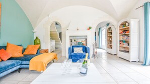 A spacious living room with a blue sofa, orange cushions, a white coffee table with wine glasses, and a blue armchair. A staircase leads to a dining area, and a bookshelf is visible in the background. - Mas des Chênes Verts – 5 ch avec piscine et clim (Eygalières)
