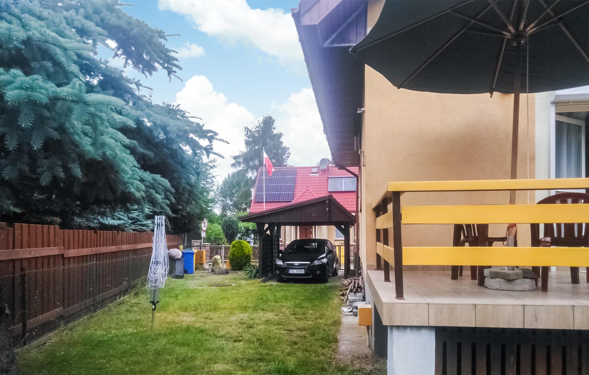 Un patio trasero con un coche estacionado bajo un porche, un área cercada y un edificio con techo rojo.
