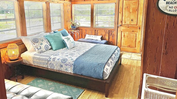 Een slaapkamer in een houten cabin-stijl met een bed, twee ramen, een plafondventilator en een bordje met de tekst 'WELCOME TO BEACH'.