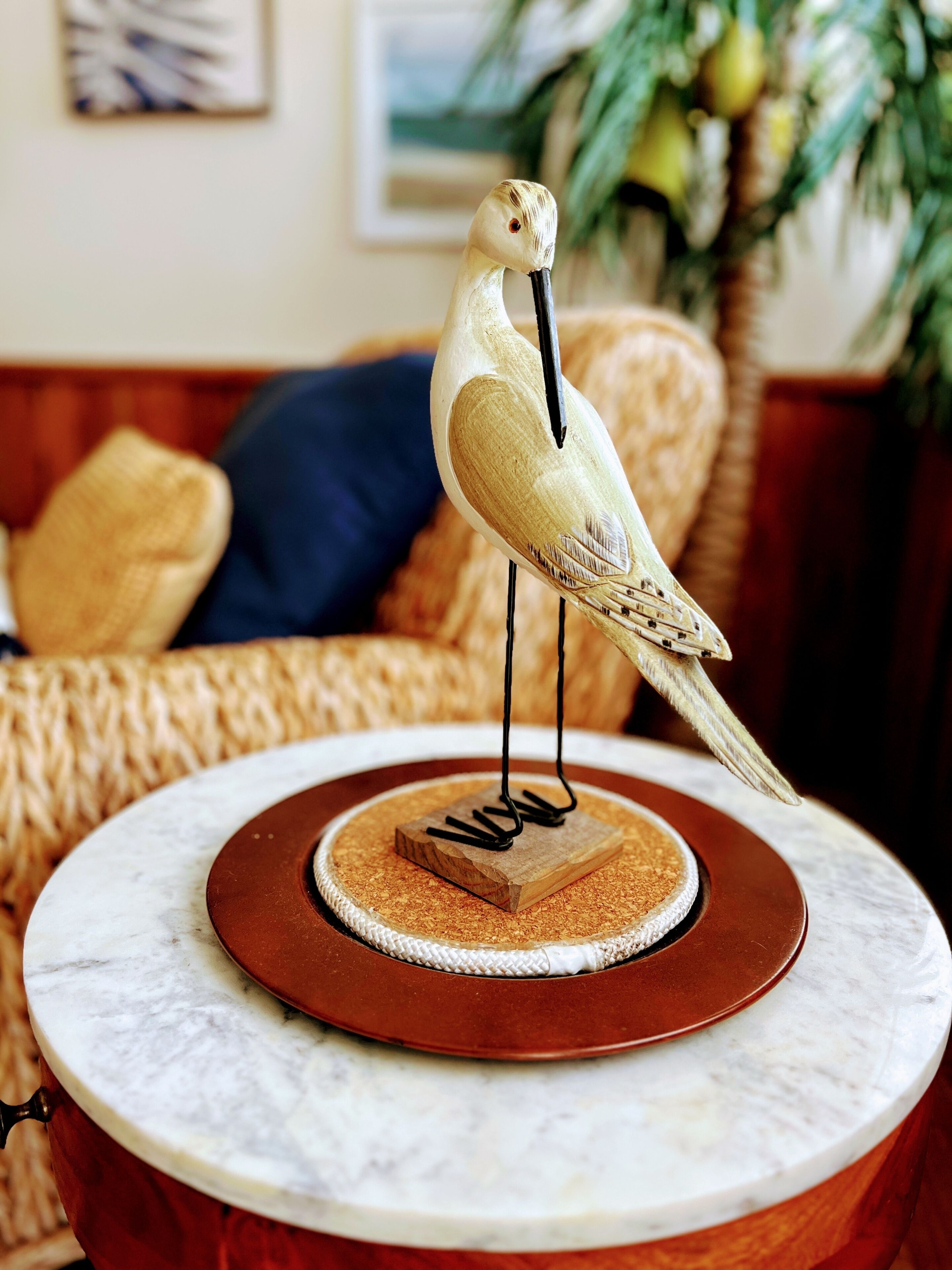 Une sculpture d’oiseau en bois est posée sur une table en marbre dont le socle est de couleur marron. La table est ronde et se trouve dans une pièce où l’on aperçoit un canapé et une plante en arrière-plan.
