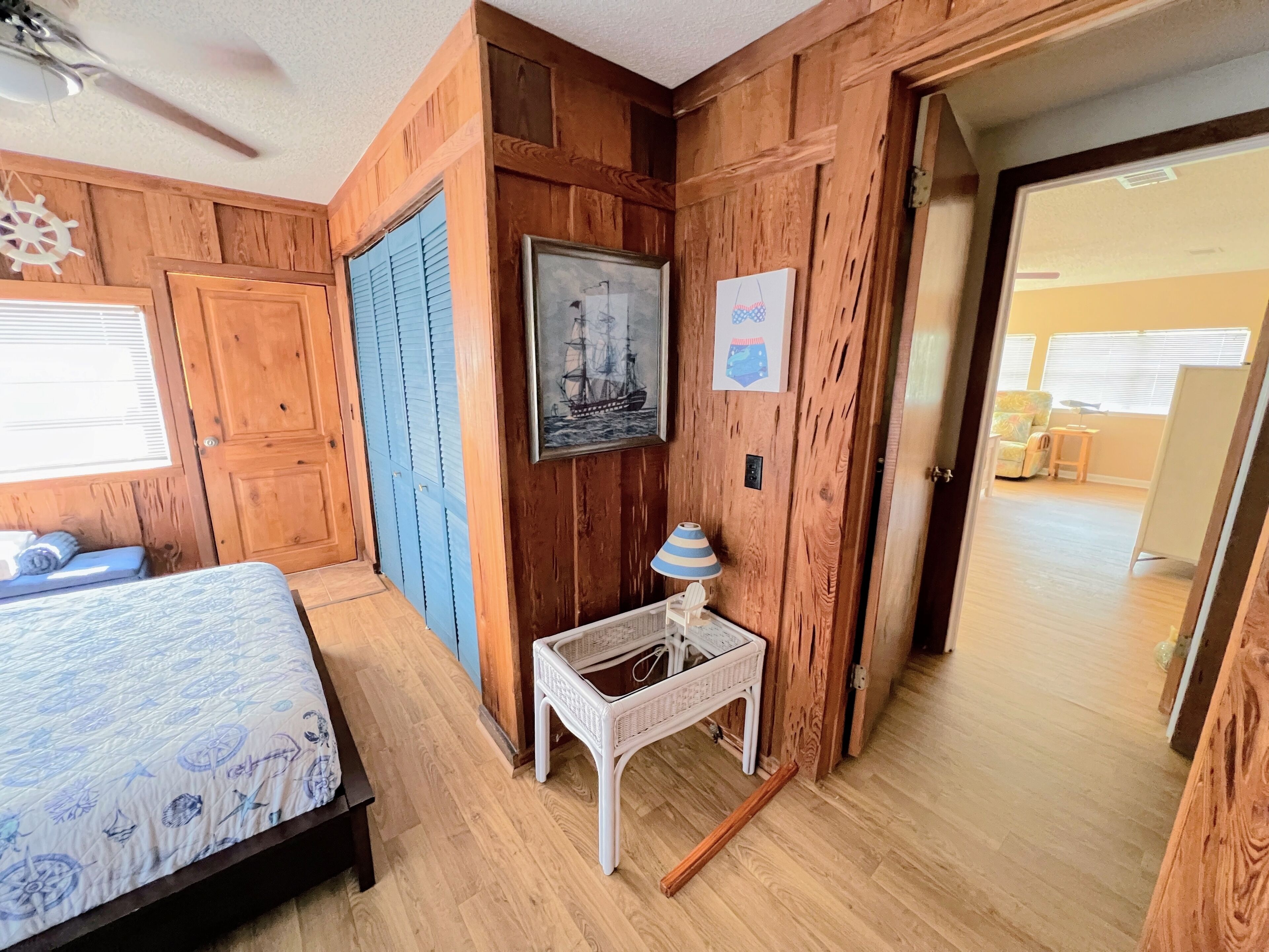A room with wooden walls, a bed with a patterned bedspread, a small white table with a lamp, and a framed picture on the wall.