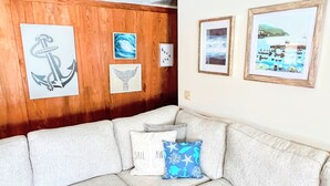 Een woonkamer met een houten wand, een beige hoekbank en drie ingelijste kunstwerken met afbeeldingen van zeedieren.