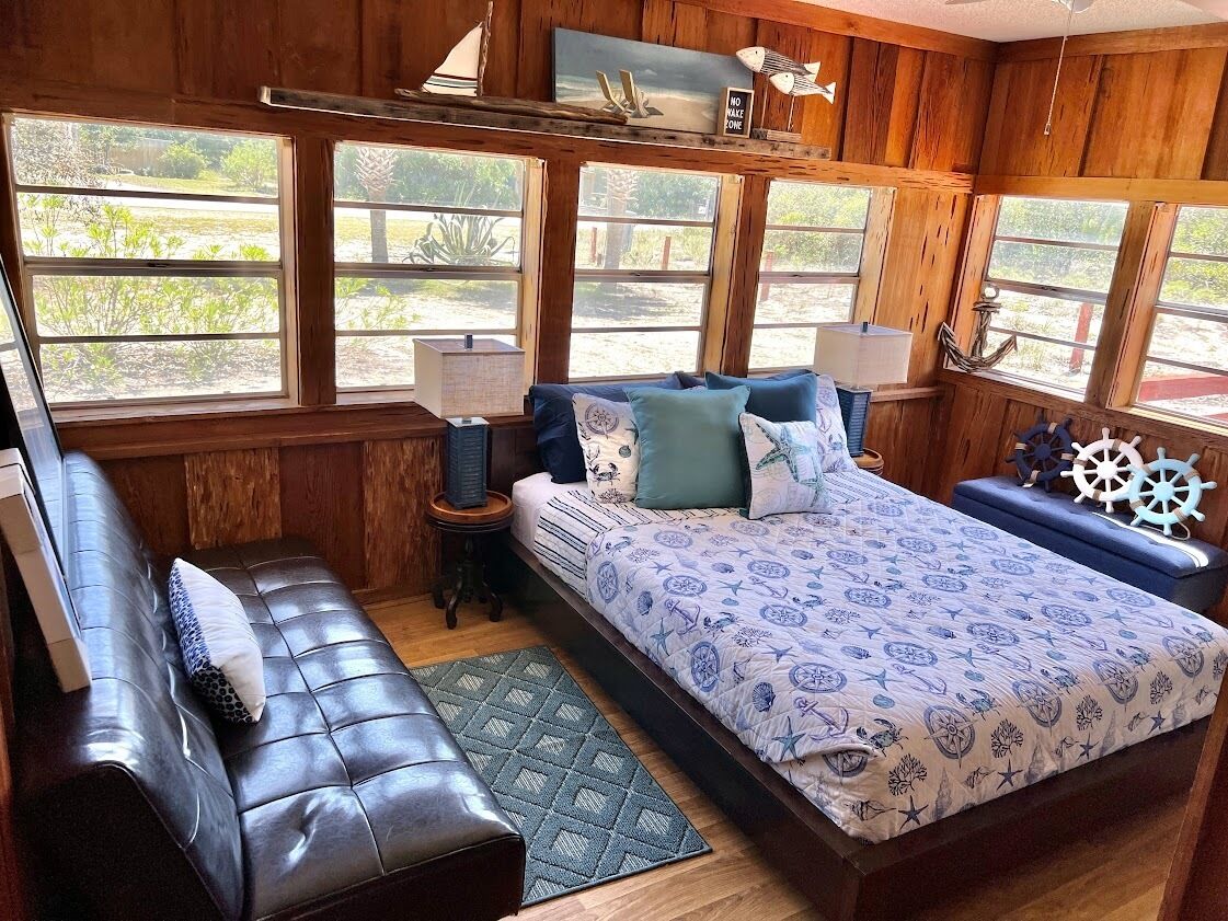 A room with a large bed, a sofa, and a desk. There are windows, a ceiling fan, and a ship wheel decoration.