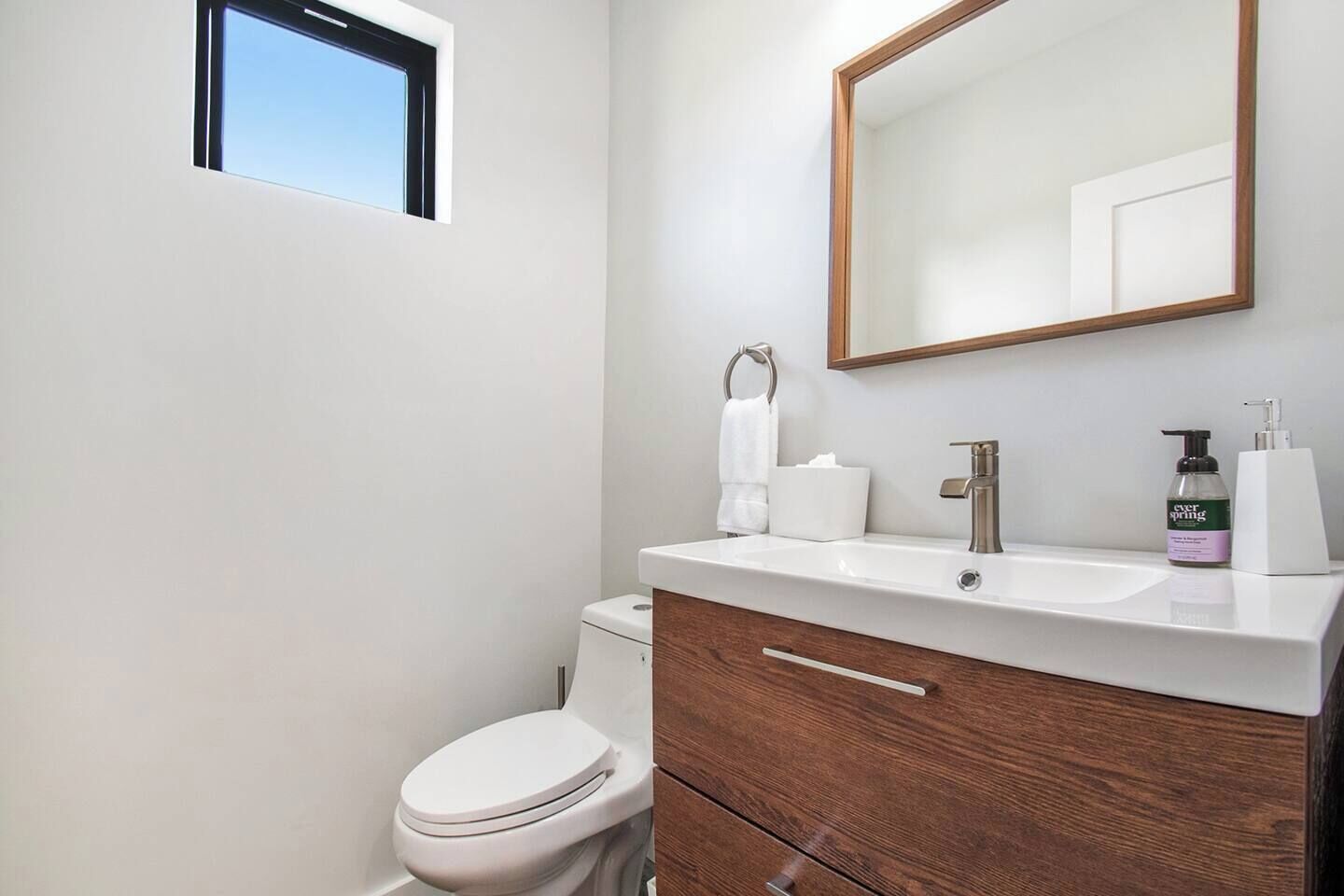 A modern bathroom with a white toilet, a wooden vanity with a mirror, and a window above the toilet.