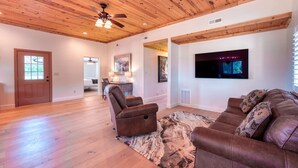 Un salon spacieux doté d’un plancher en bois, d’un ventilateur de plafond, d’un grand téléviseur et d’un canapé en cuir marron.