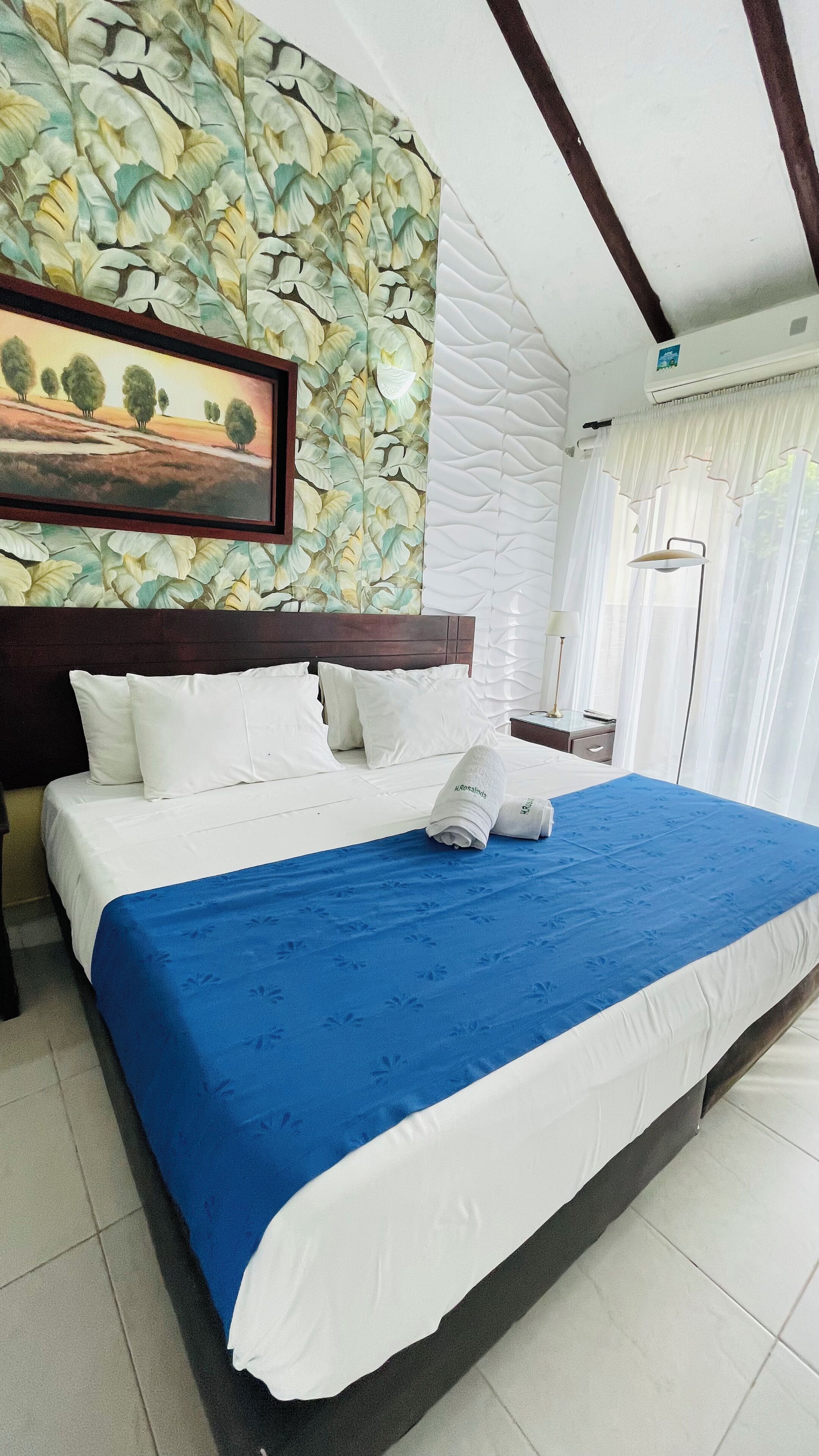 Una cama bien hecha con una colcha azul y blanca, almohadas blancas y un cuadro enmarcado de un paisaje en la pared de arriba.