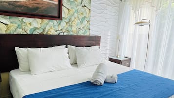 Una cama bien hecha con una colcha azul y blanca, almohadas blancas y un cuadro enmarcado de un paisaje en la pared de arriba.