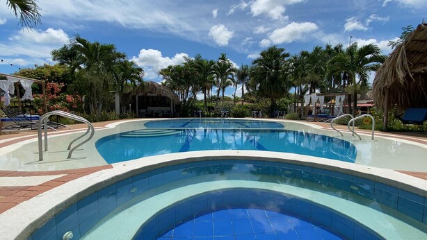 A swimming pool with a curved edge, surrounded by palm trees and lounge chairs. - Hotel Rosalinda Campestre del Llano (Villavicencio)