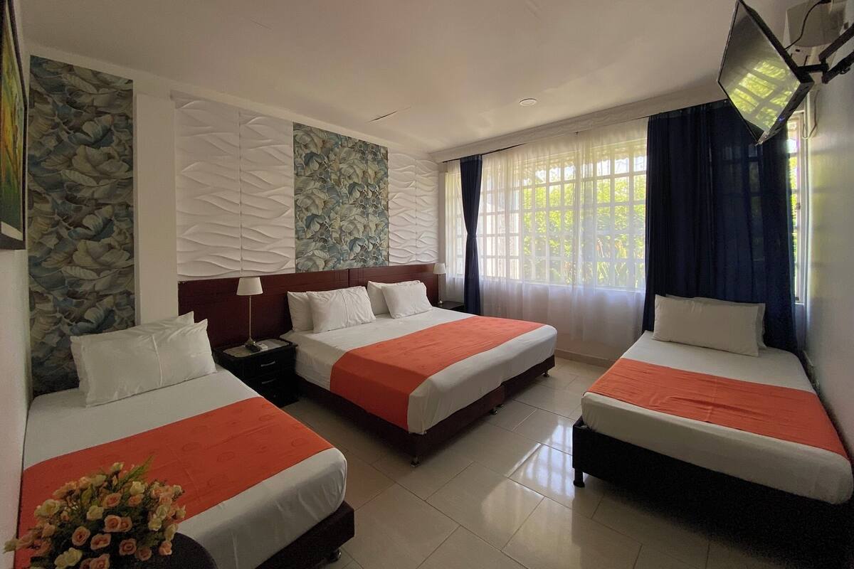 A hotel room with three single beds, a stone accent wall, a large window with curtains, and a flat-screen TV mounted on the wall.