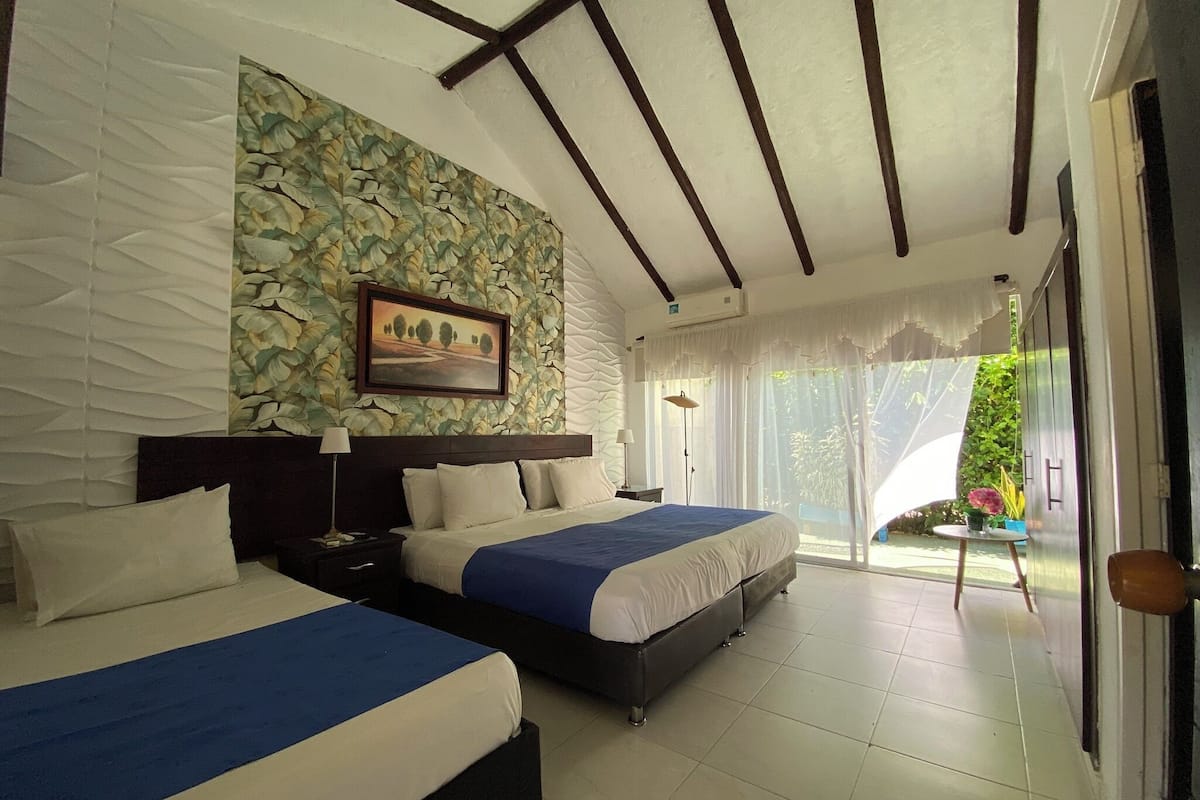 A hotel room with two beds, a stone wall with a floral pattern, a framed picture, and a view of greenery outside.
