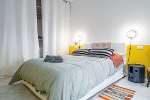 A bed with a grey and white striped pillow, a grey blanket, and a grey towel on the floor. There is a small round table with a lamp, a trash bin, and a yellow wall.