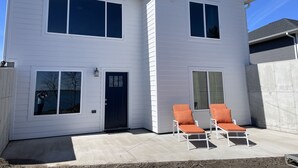A two-story house with a dark front door, large windows, and a concrete patio with two lounge chairs.
