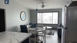A modern kitchen with a marble countertop, a dining area with a round table and chairs, a ceiling fan, and a large window with a view of trees.